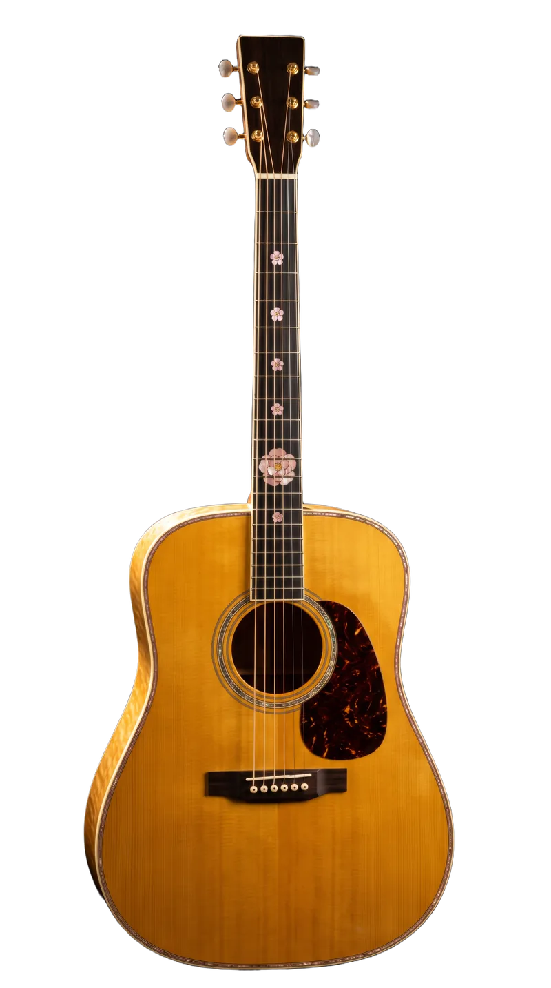 A butterscotch dreadnought acoustic with a tortoiseshell pickguard and figured sides, standing upright against pure black, small pink-pearl camellias inlaid down its fingerboard to a full bloom at the twelfth fret.